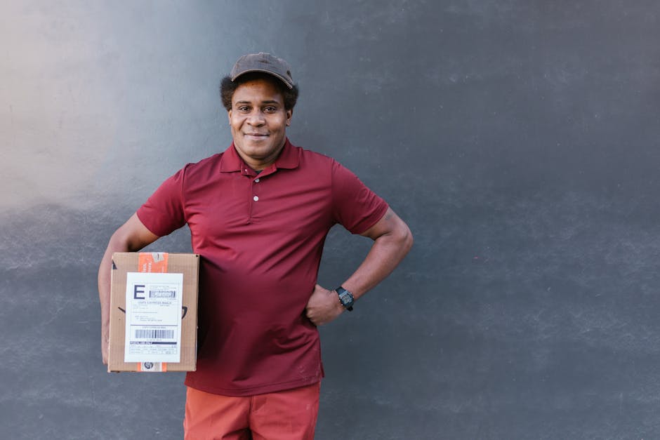 A male flower delivery courier with medium brown skin and curly black hair, wearing a maroon polo shirt, matching maroon trousers, and a grey cap, stands against a plain dark grey wall. He holds a med